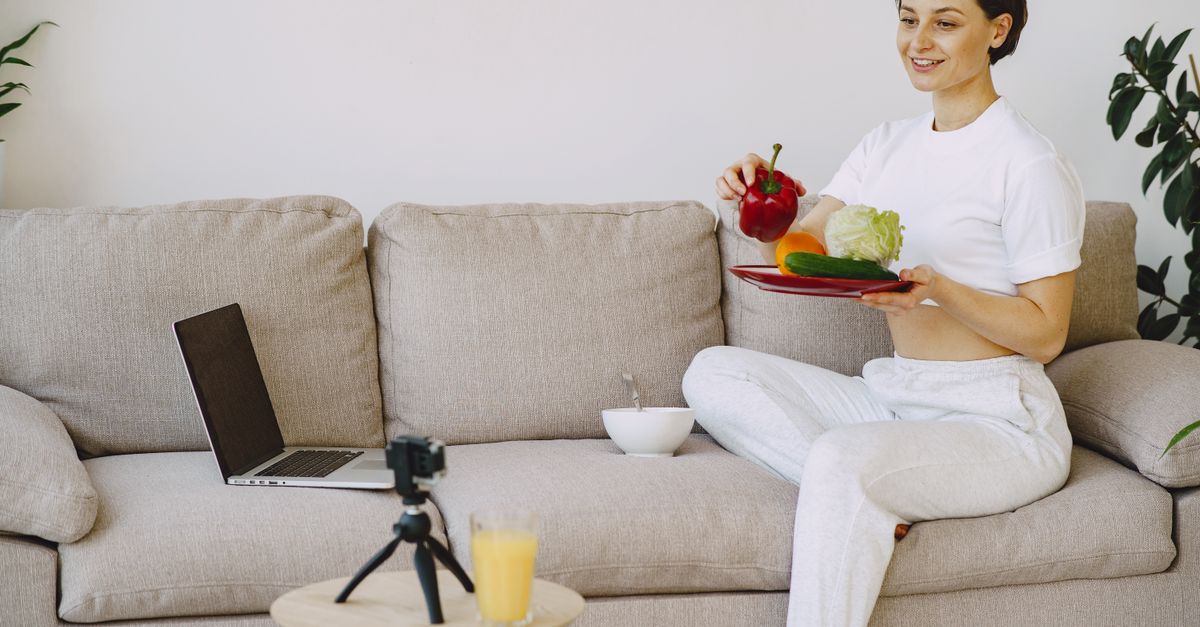 Content female in white outfit sitting on sofa holding plate with healthy food in hand and taking photo on camera
